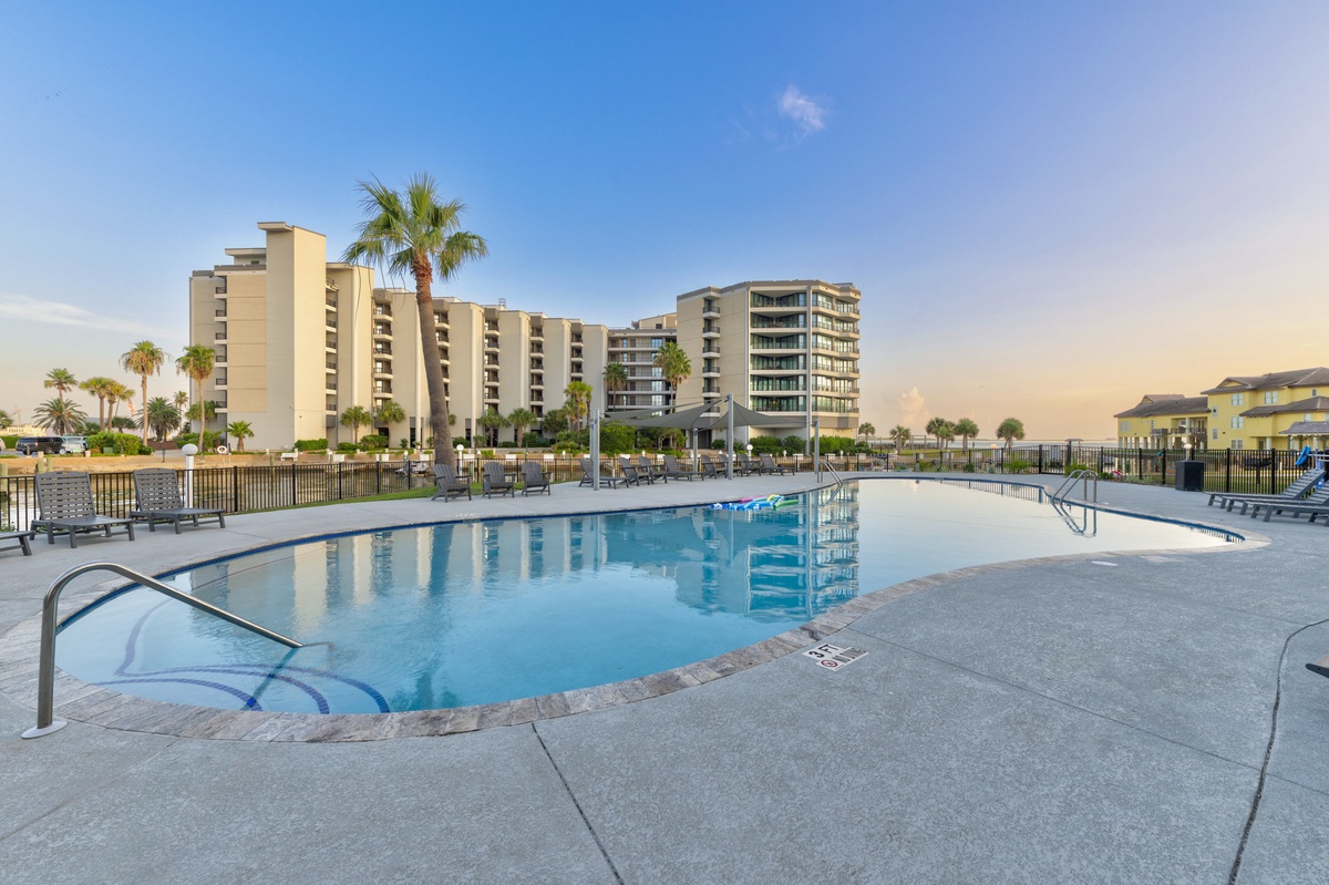 A spacious outdoor pool surrounded by lounge chairs and tropical palms, set within a beachfront resort complex under clear skies.