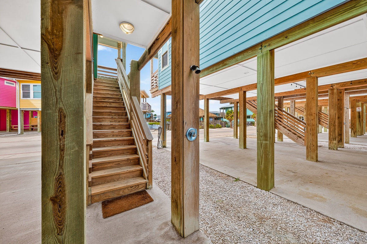 This charming stilt-house entrance leads to a bright coastal escape with weathered wood beams, a cheerful blue exterior, and colorful neighbors in view. A welcoming space that captures the relaxed spirit of beachside living.