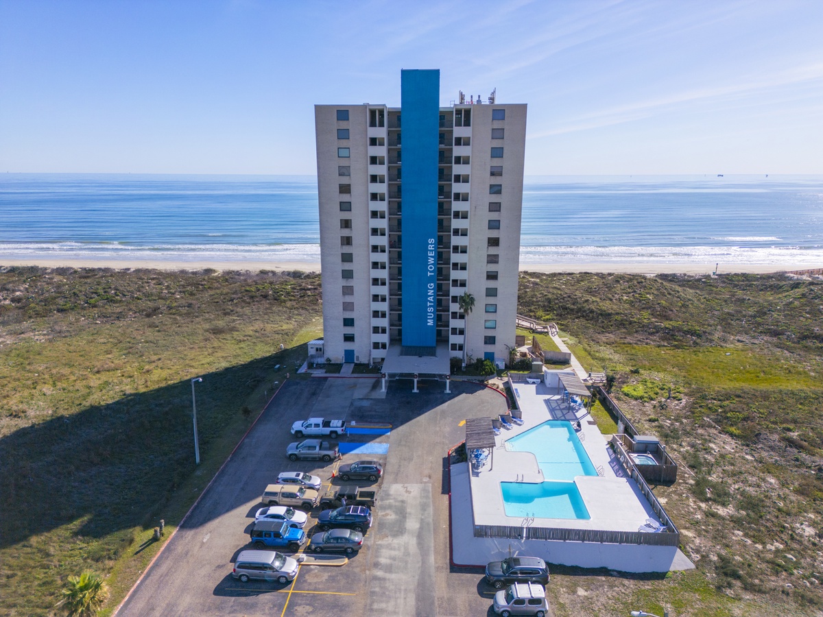 Oceanfront high-rise tower with pool and parking, surrounded by coastal vegetation near pristine beach.