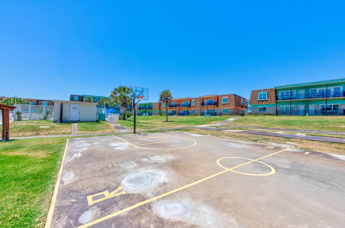 Outdoor basketball court with full hoops and painted lines, surrounded by colorful resort buildings under clear blue skies.