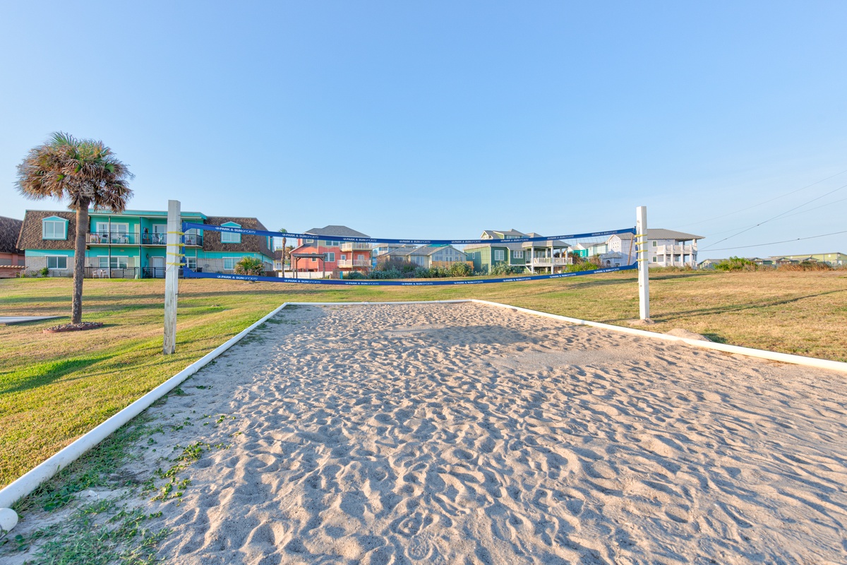 Beach volleyball court with net and sand surrounded by colorful coastal homes under clear blue skies.