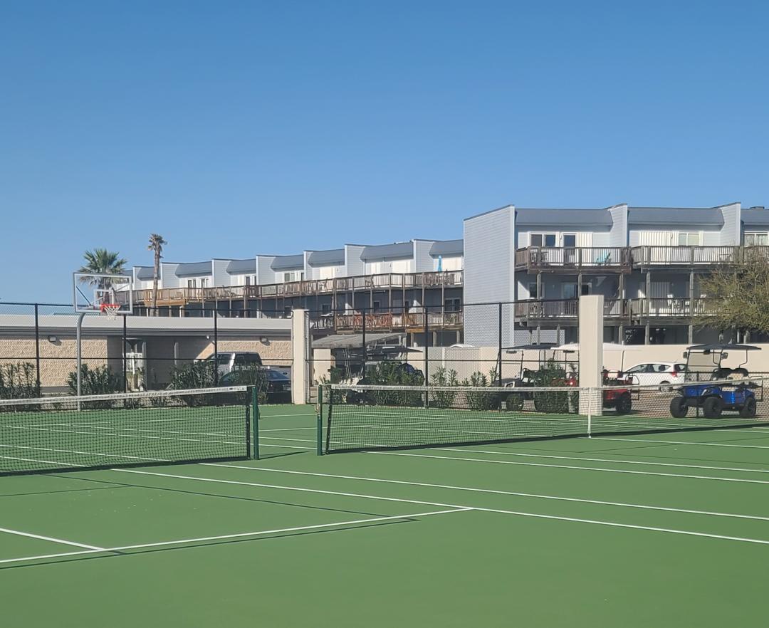 A well-maintained tennis court with property buildings and parking visible in the background under clear blue skies.