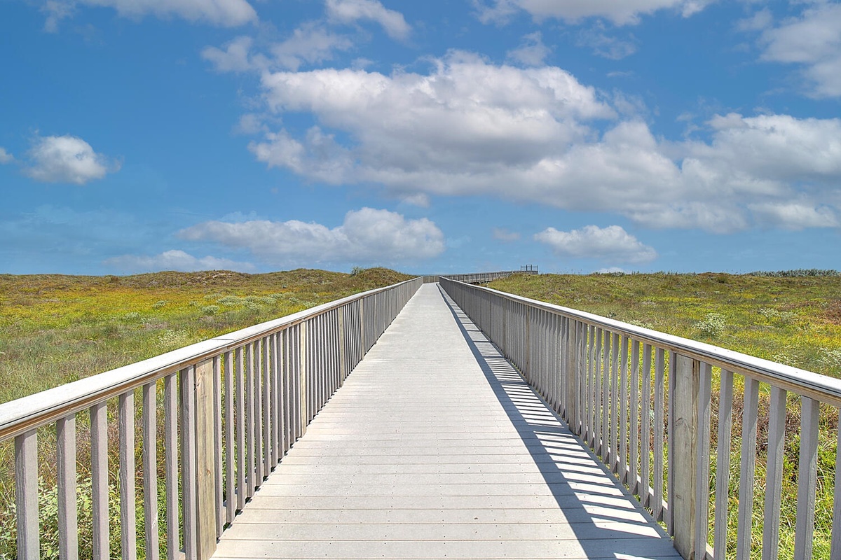 Wooden boardwalk stretches across coastal dunes toward the horizon under blue skies with scattered clouds.