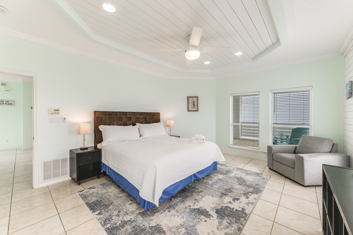 Relax in your bright, coastal-inspired bedroom featuring a plush king bed, cozy seating area, and ceiling fan for ultimate comfort.