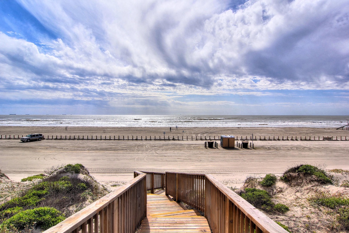 Dramatic sky formations sweep over pristine coastline where wooden boardwalk leads to expansive sandy beach and rolling waves.