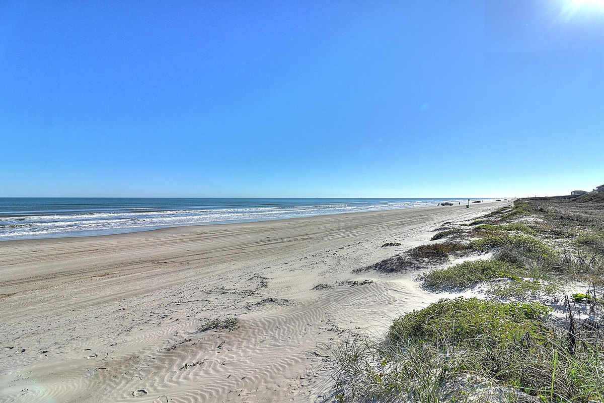 Pristine sandy coastline stretches endlessly under brilliant blue skies, with gentle waves and natural dunes creating the perfect beachside setting.