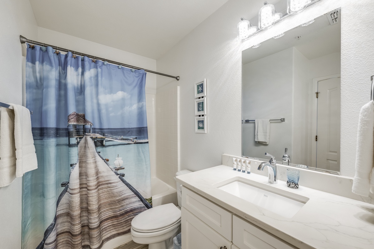 Step into a fresh, serene bathroom with gleaming countertops, soft white towels, and a scenic tropical shower curtain that brings vacation vibes to your morning routine.