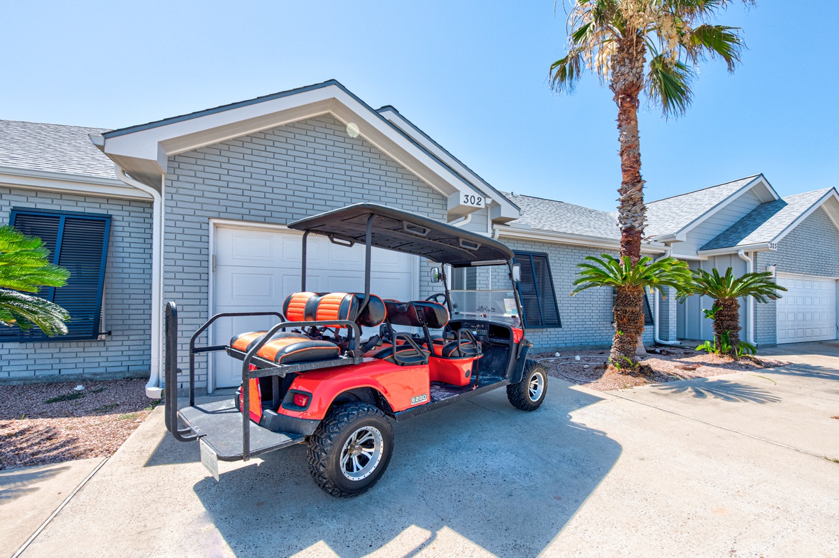 Modern vacation rental with golf cart parked in covered driveway, surrounded by palm trees and tropical landscaping in sunny residential area.
