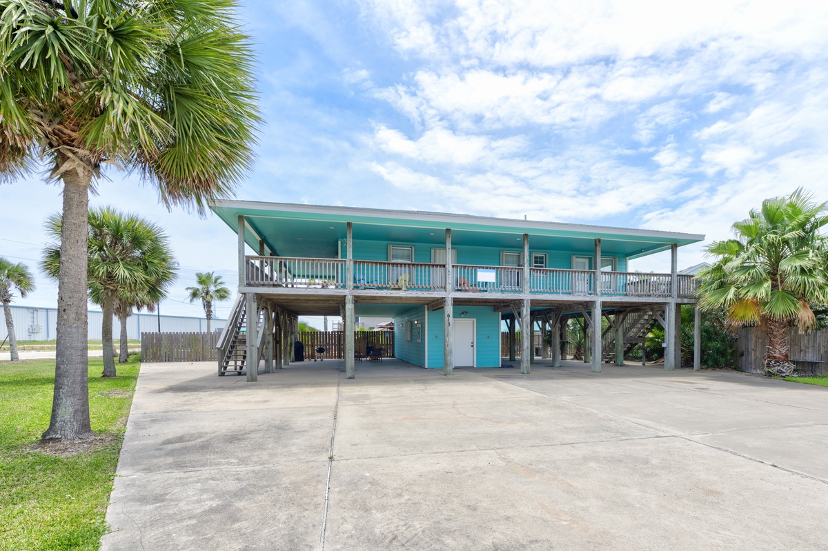 A distinctive turquoise beach house elevated on stilts, surrounded by swaying palms and open coastal land, offering a classic coastal retreat experience.