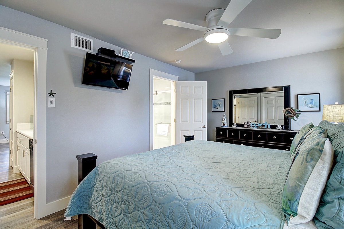 Drift off in your serene master suite with cool ocean-hued linens, ceiling fan comfort, and wall-mounted TV for cozy evening entertainment.