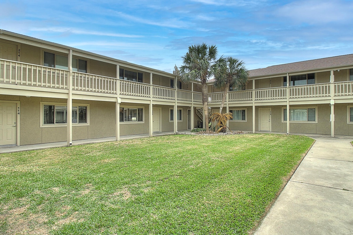 Two-story property buildings frame a peaceful central courtyard with lush green lawn and tropical palm trees under bright blue skies.