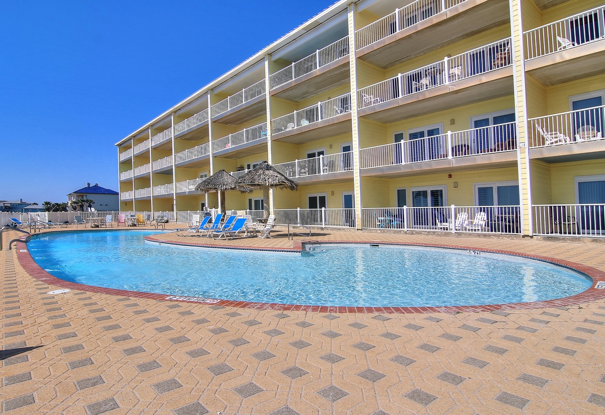 Multi-story beachfront property featuring an outdoor swimming pool with sun loungers and tiki umbrellas, set against clear blue skies.