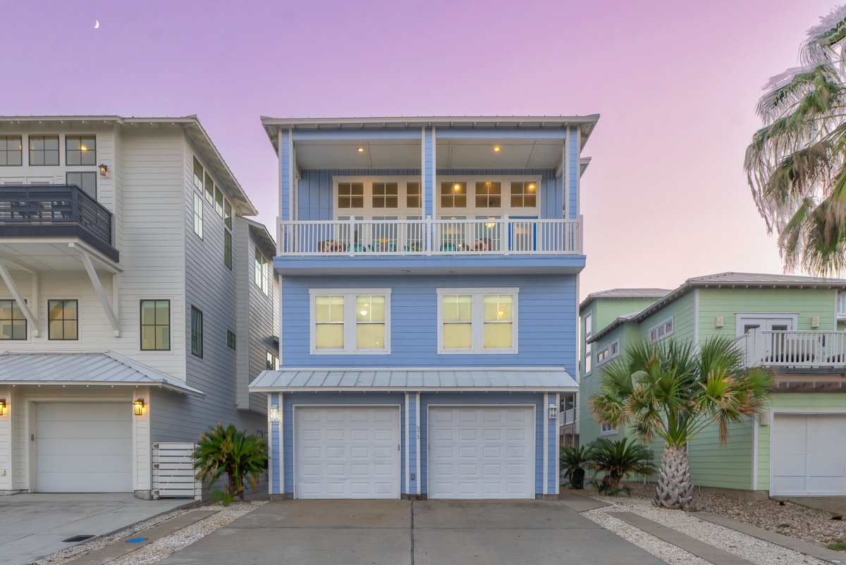 Coastal beach house with tropical landscaping and colorful neighboring properties under a beautiful purple sunset sky.