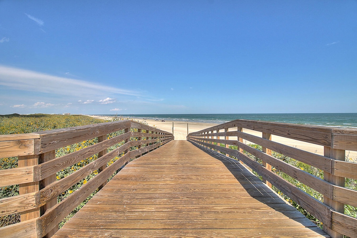Wooden boardwalk leads across coastal dunes to pristine beach where ocean meets sandy shore under brilliant blue skies.