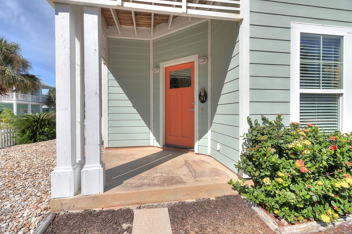 A welcoming coastal cottage entrance with sage green siding and cheerful coral door, framed by lush tropical landscaping and white pillars.