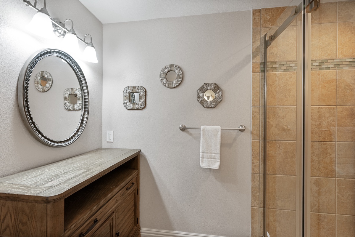 Step into your beautifully appointed bathroom featuring elegant mirrors, warm wood vanity, and spacious glass shower with travertine tile.