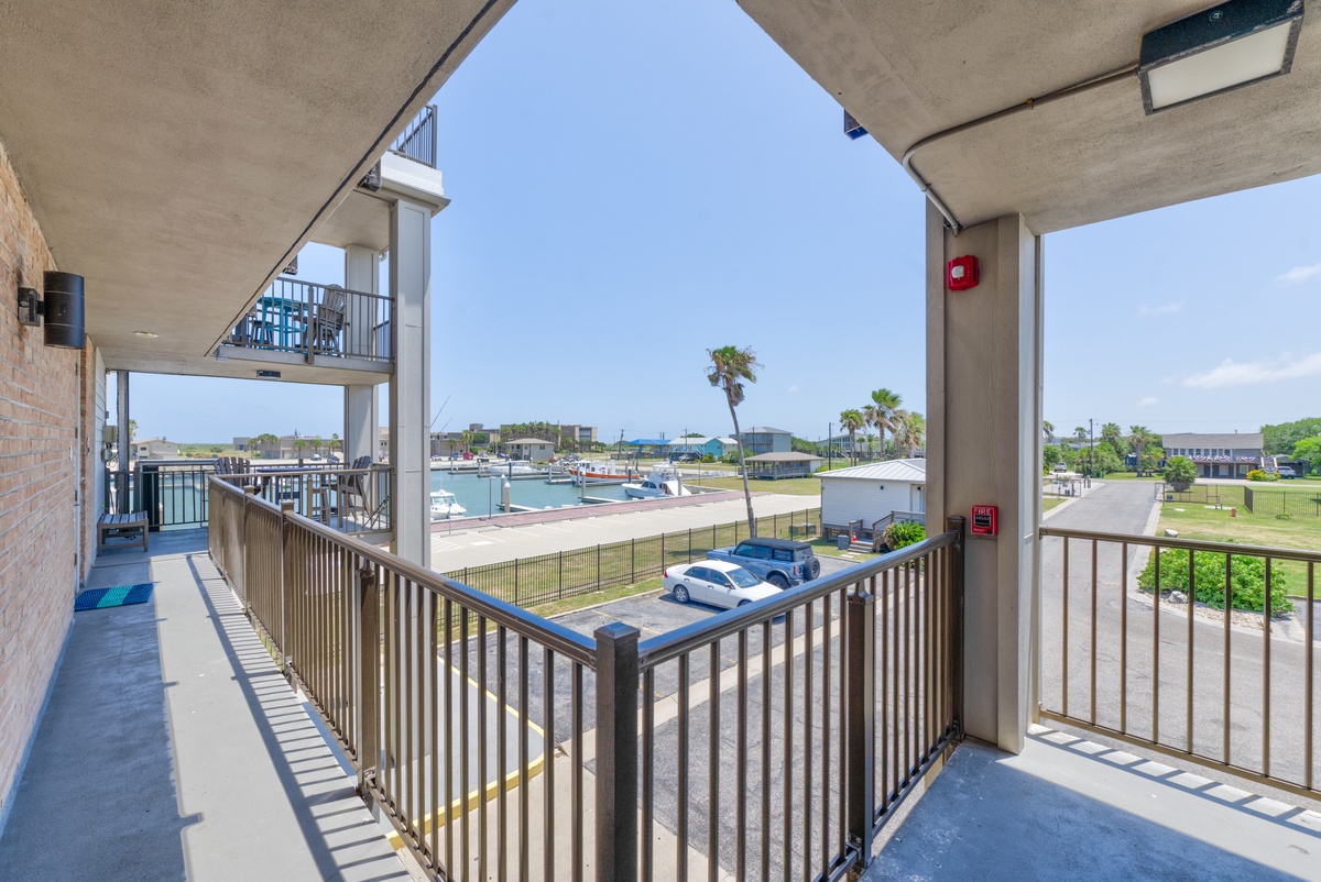 Covered balcony overlooks marina and waterfront with boats, palm trees, and coastal buildings visible in the distance.