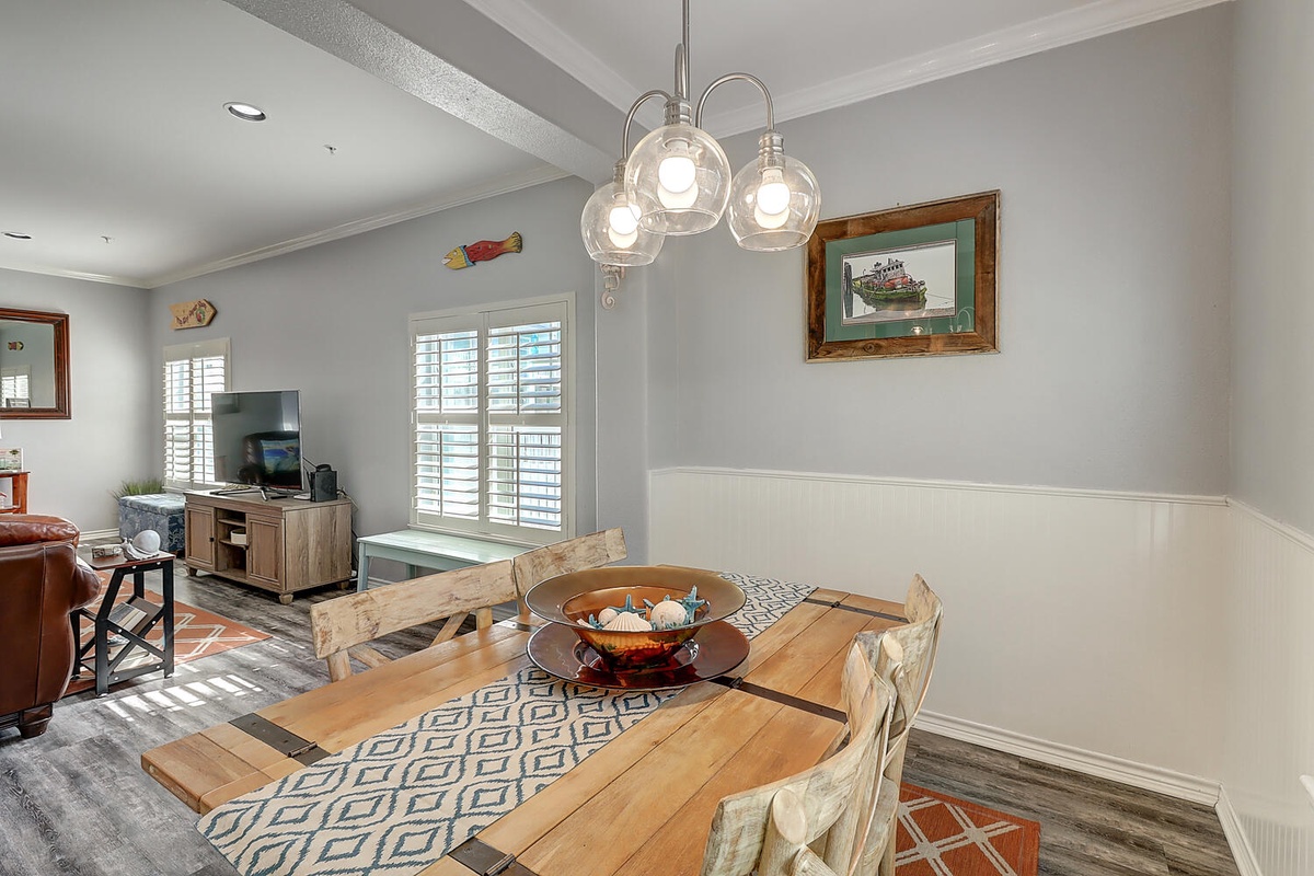 Your bright dining space features coastal charm with a rustic wooden table, pendant lighting, and an open view to the living area.