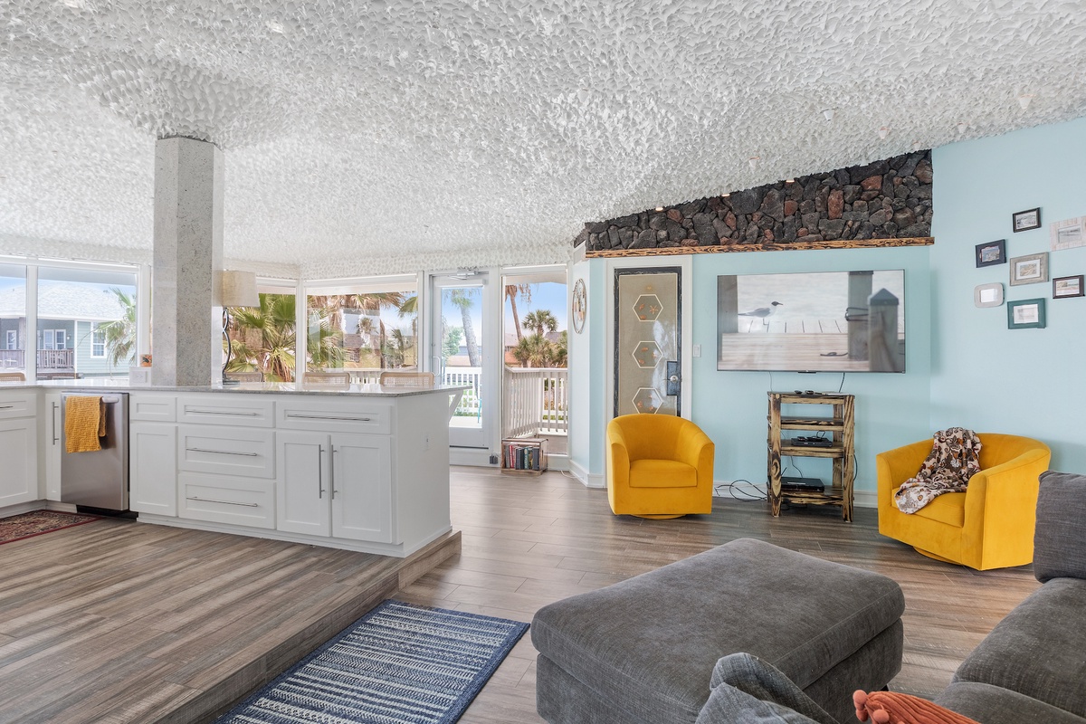 Relax in your bright open living space with vibrant yellow accents, modern kitchen, and windows framing tropical palm views.