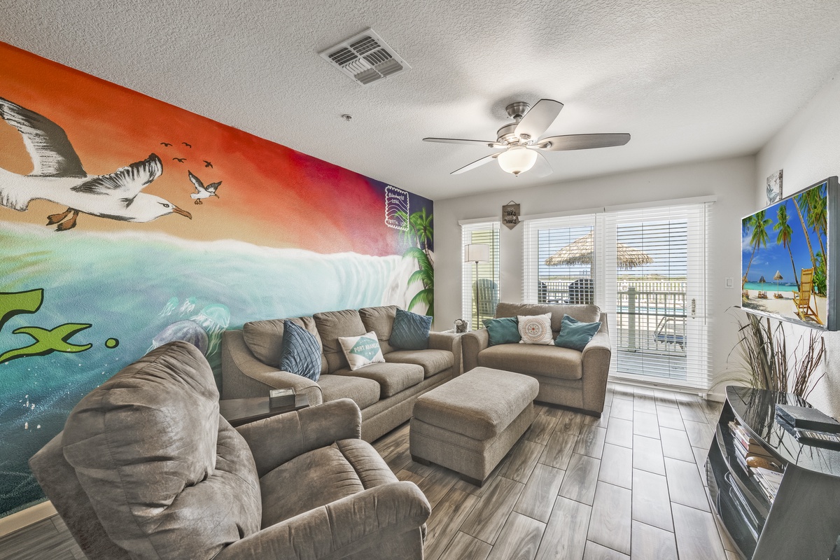Relax in your bright coastal-inspired living room with plush seating, a vibrant ocean mural, ceiling fan, and doors opening to your private balcony.