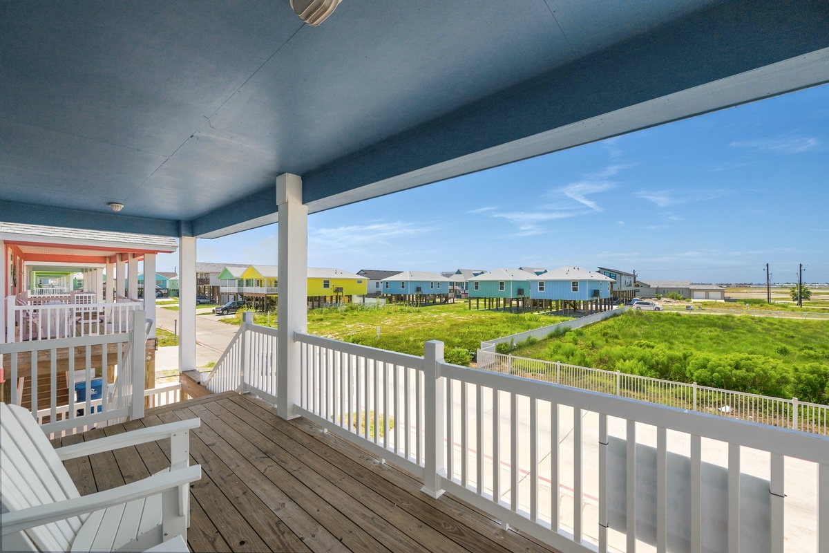 A covered deck with Adirondack chairs frames a view of colorful beachfront neighborhood homes, open green space, and expansive coastal scenery.