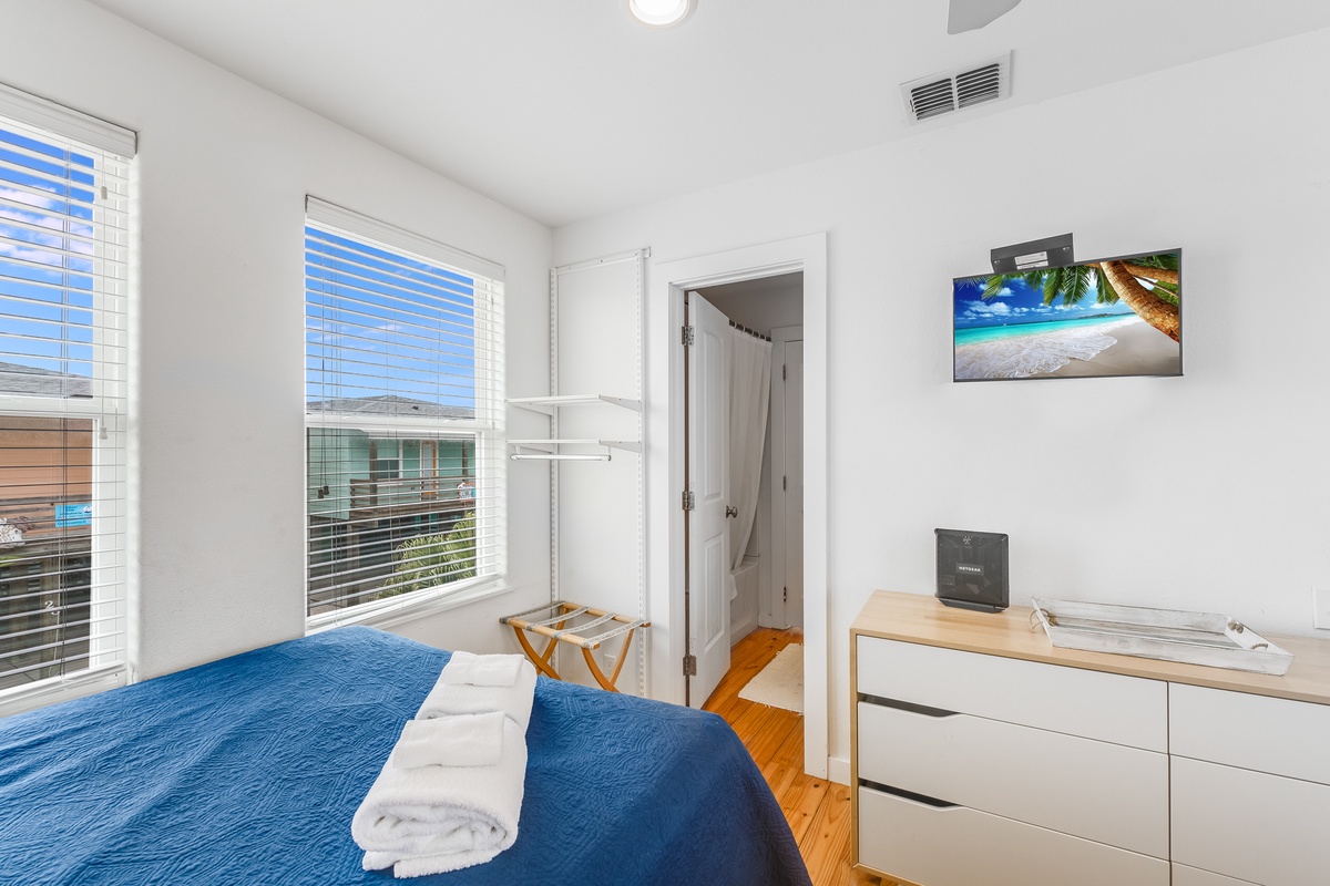 Relax in your bright, airy bedroom with fresh blue linens, modern amenities, and views of the neighborhood through white blinds.
