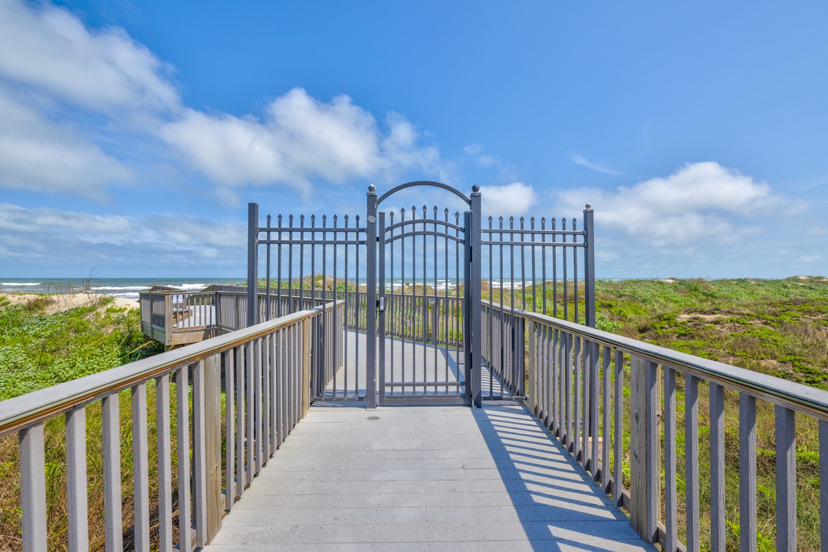 A pedestrian boardwalk leads through coastal dunes to the sandy beach beyond, with ocean waves visible under a partly cloudy blue sky.