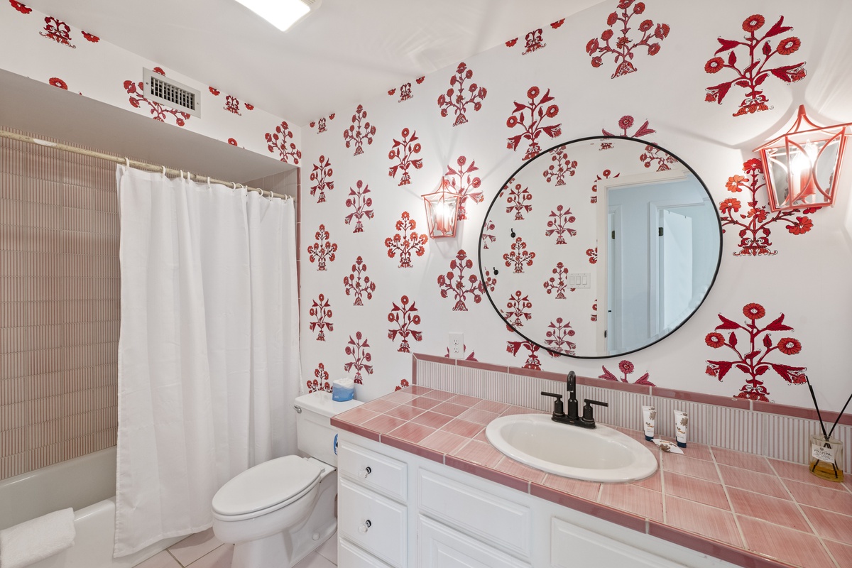 Your charming bathroom features vibrant red floral wallpaper, pink tile counters, and warm lighting—creating a cozy, stylish space to freshen up.