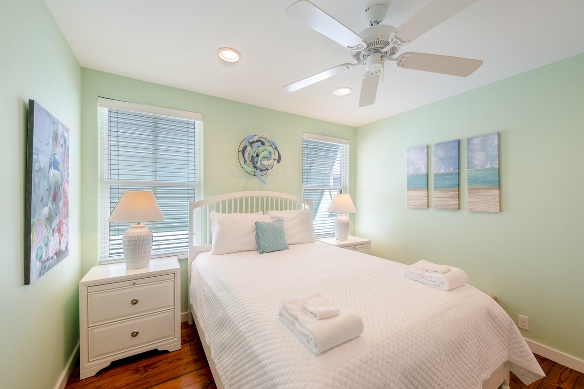 Drift off in your serene coastal bedroom with soothing green walls, crisp linens, and beach-themed artwork creating perfect vacation vibes.