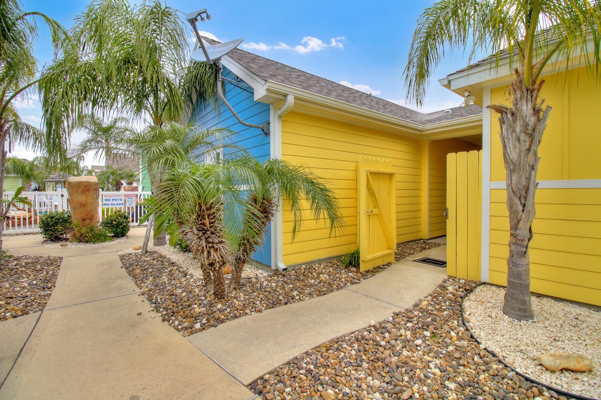 Bright tropical cottage with vibrant yellow and blue exterior, surrounded by swaying palm trees in a sunny beachside setting.