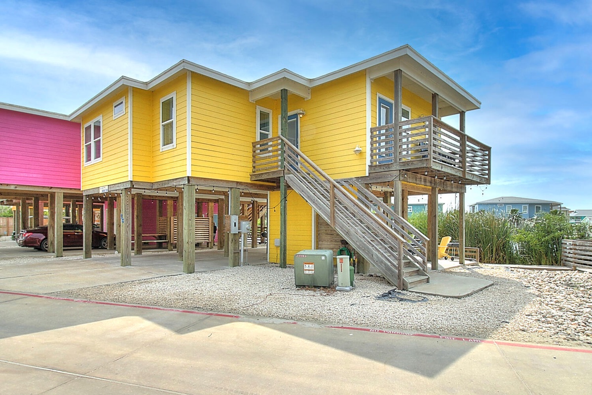 Bright yellow elevated beach house with covered parking and wooden stairs leading to the main entrance.