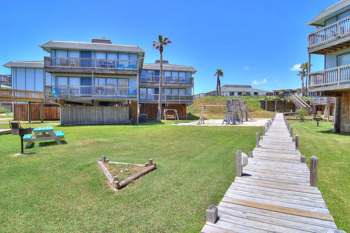 Beachfront property with wooden walkway leading to multi-level vacation rental buildings and playground area.
