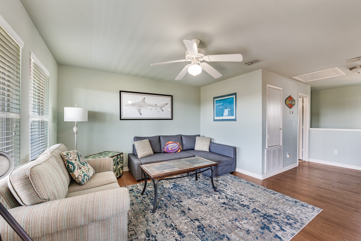 Relax in your spacious living room with comfortable seating, coastal artwork, and plenty of natural light streaming through plantation shutters.