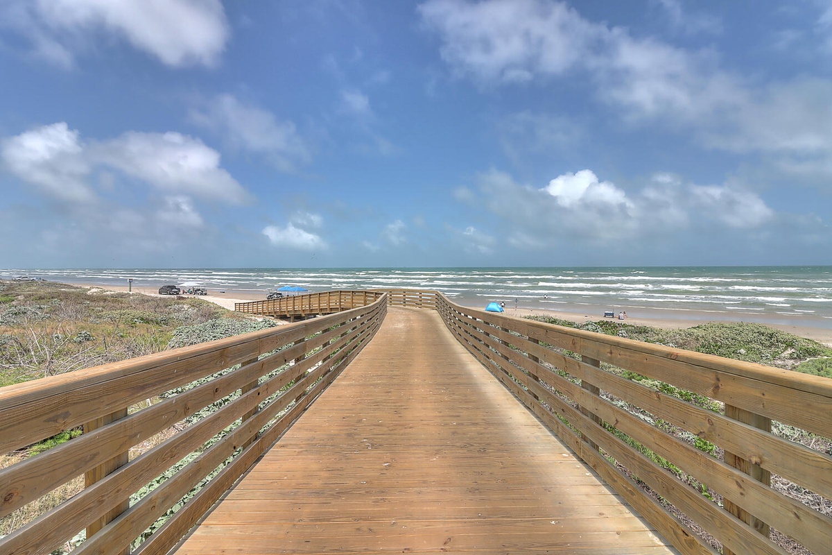 A wooden boardwalk extends toward pristine sandy shores where ocean waves meet the coastline under expansive blue skies.