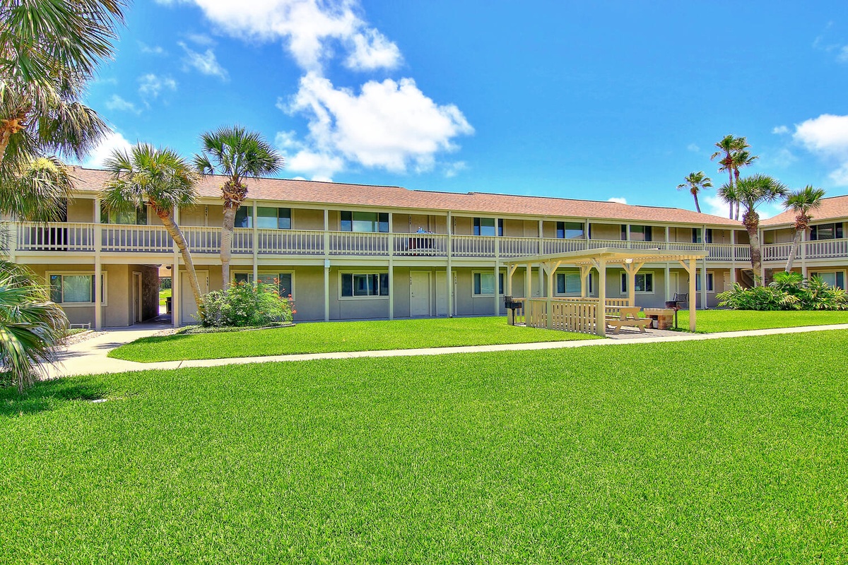 Two-story property building featuring tropical landscaping with palm trees and expansive green lawn areas under sunny Florida skies.