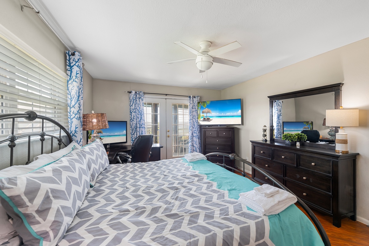 Your coastal-inspired bedroom retreat features a comfortable queen bed, ceiling fan, and double doors opening to your private balcony. Fresh towels await on your bed as soft light filters through airy curtains.