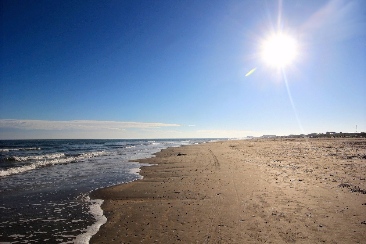Pristine sandy beach stretches along sparkling ocean waters under brilliant sunshine, with gentle waves lapping the shore.