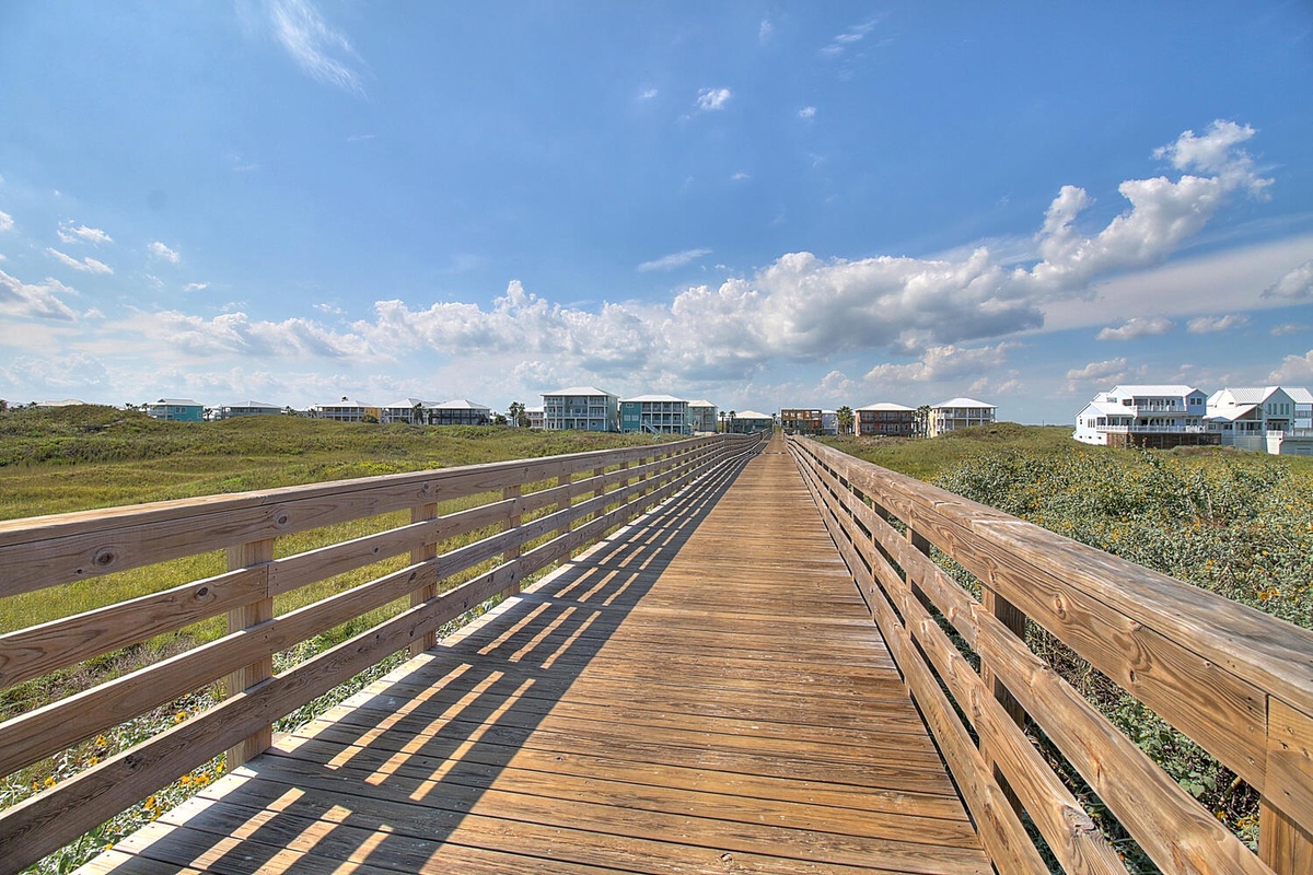 Wooden boardwalk leads through coastal landscape toward beachfront vacation homes under expansive blue sky with scattered white clouds.
