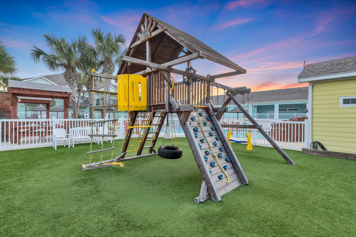 Vibrant playground with climbing features and swings set against a stunning sunset backdrop, surrounded by colorful vacation homes.