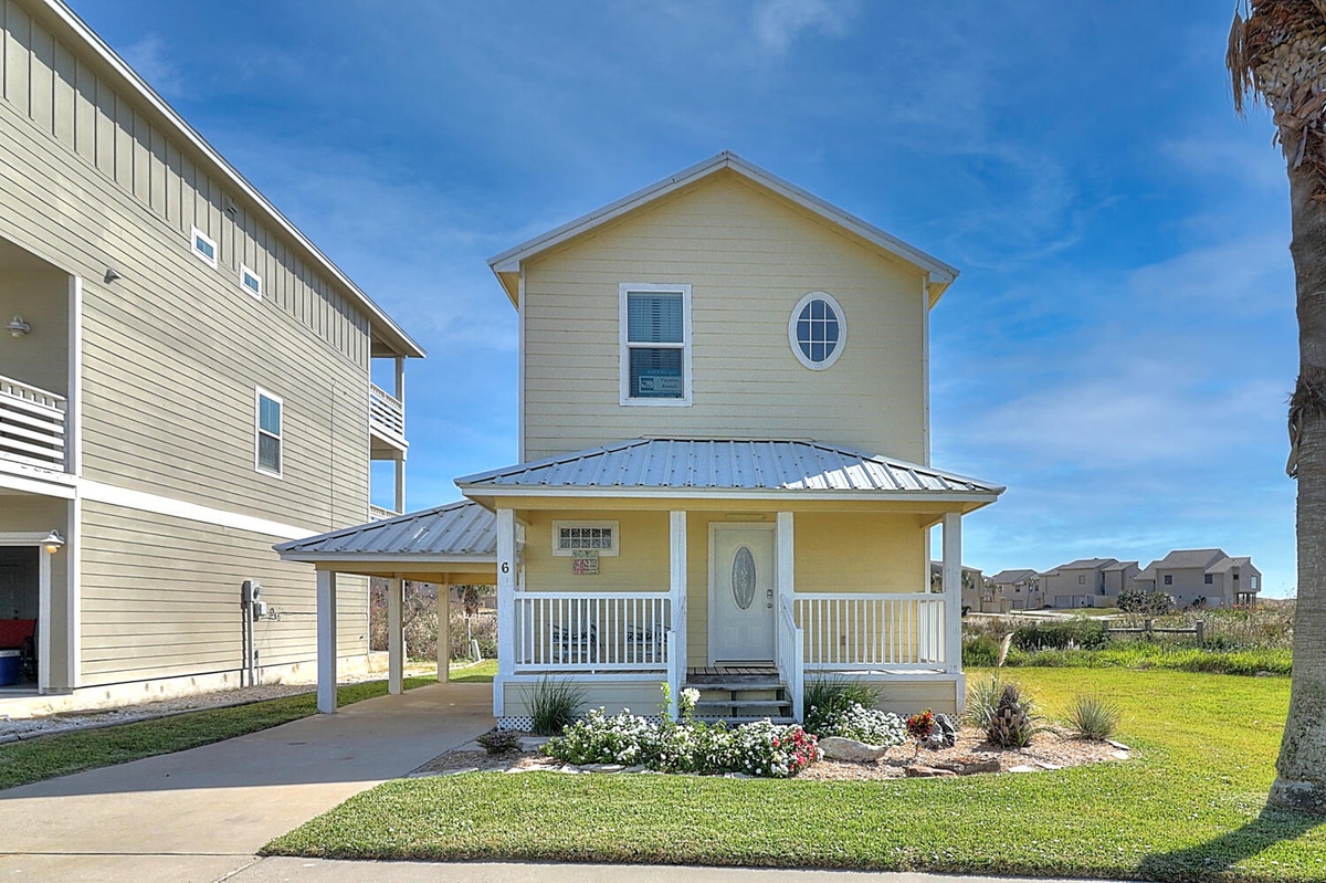 Bright yellow beach house with white trim and wraparound porch, surrounded by manicured landscaping in a coastal neighborhood.