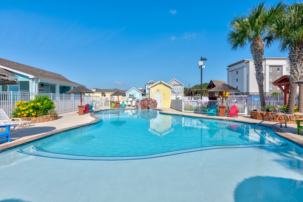 Sparkling pool complex surrounded by colorful beach cottages and palm trees offers endless summer relaxation under clear blue skies.