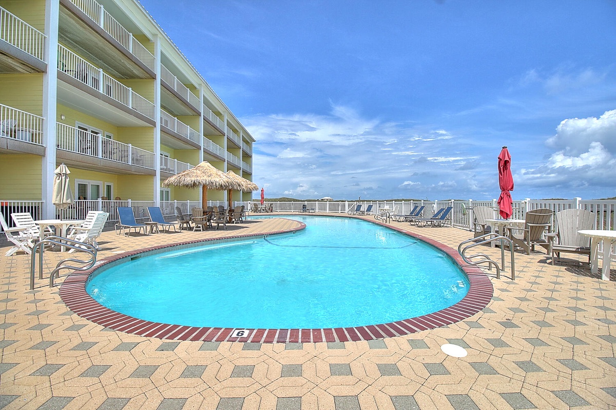 Beachfront property with expansive pool deck featuring crystal-clear swimming pool surrounded by comfortable lounge chairs under bright blue skies.