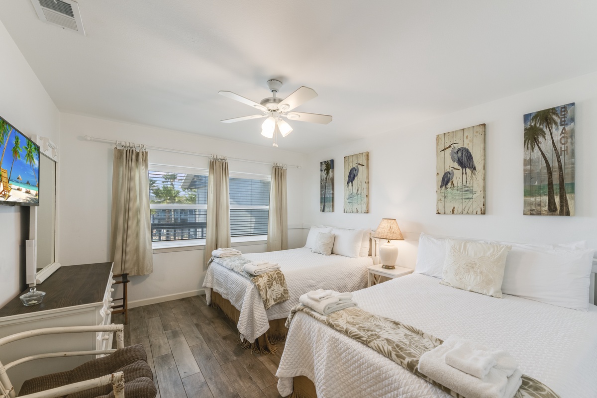 Your airy coastal bedroom features two plush beds, soft linens, and serene coastal artwork. Relax in comfort with the gentle breeze from your ceiling fan and natural light from windows.