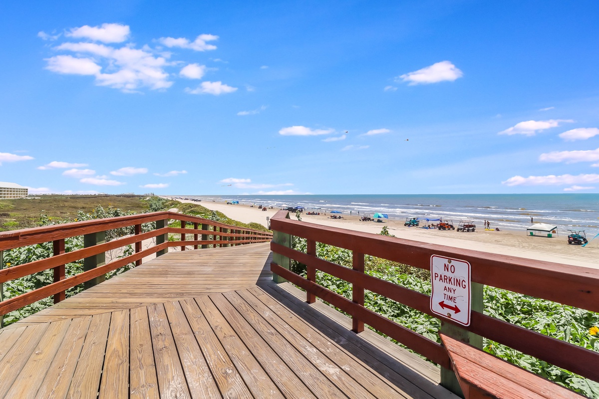 Wooden boardwalk overlooks expansive beach with vehicles driving on sand under bright blue sky with scattered clouds.