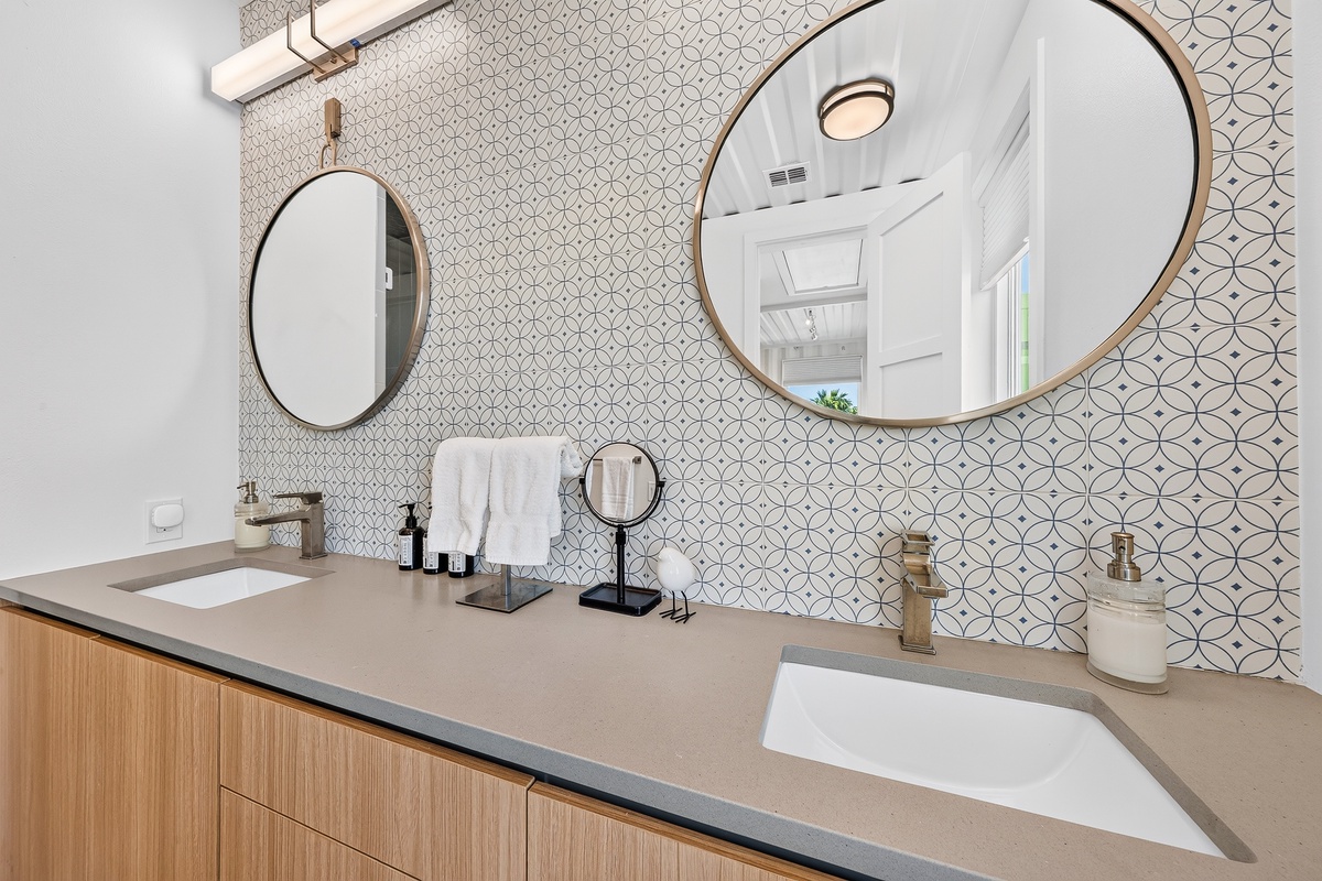 Step into your stylish dual-vanity primary bathroom where patterned mosaic tile meets modern elegance. Brass-framed mirrors catch the light while plush towels and thoughtful amenities await your arrival.