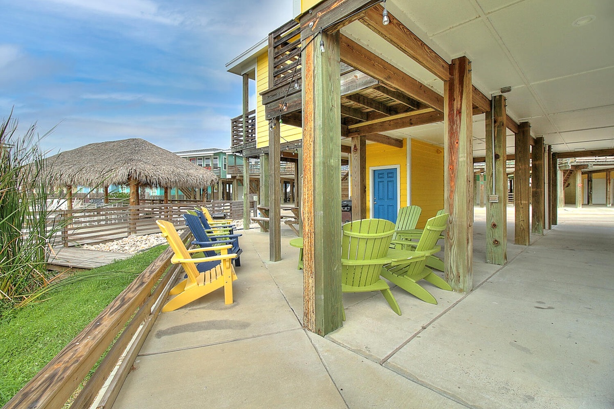 Vibrant beach house exterior with colorful Adirondack chairs under covered porch and thatched-roof pavilion nearby.