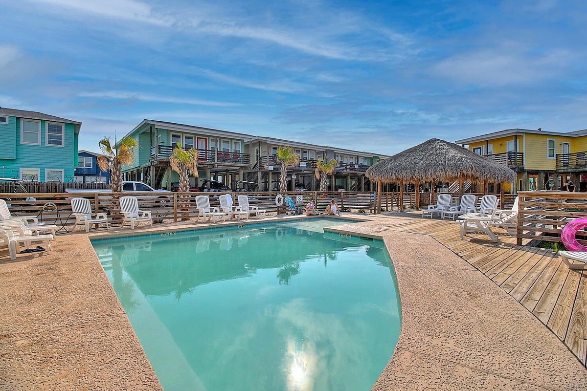 Colorful beachside resort buildings surround a refreshing pool area with tropical tiki hut and palm trees.