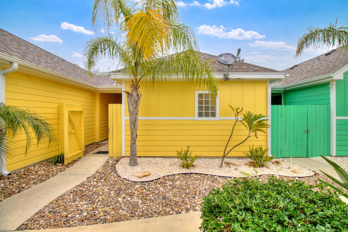 Vibrant yellow and green tropical cottages create a cheerful island-style welcome beneath swaying palm trees and bright blue skies.