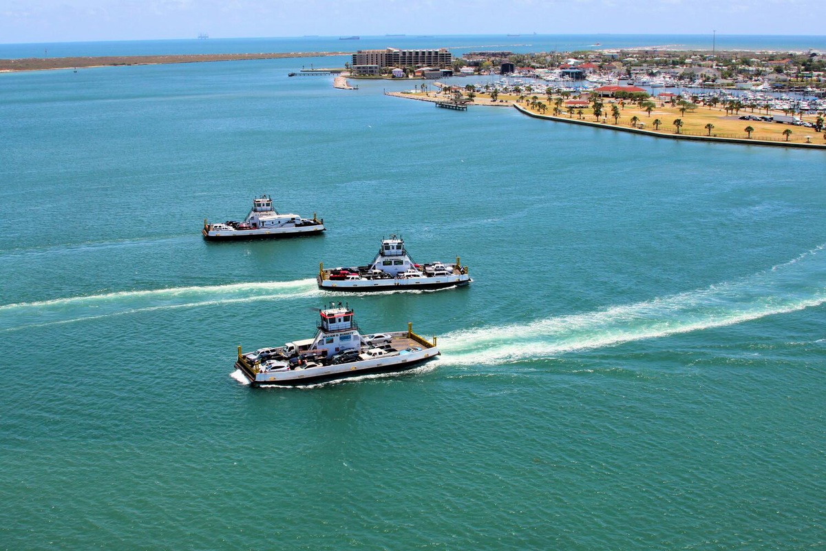 Aerial view captures three ferries navigating turquoise waters near a coastal town with palm-lined shores and waterfront buildings.
