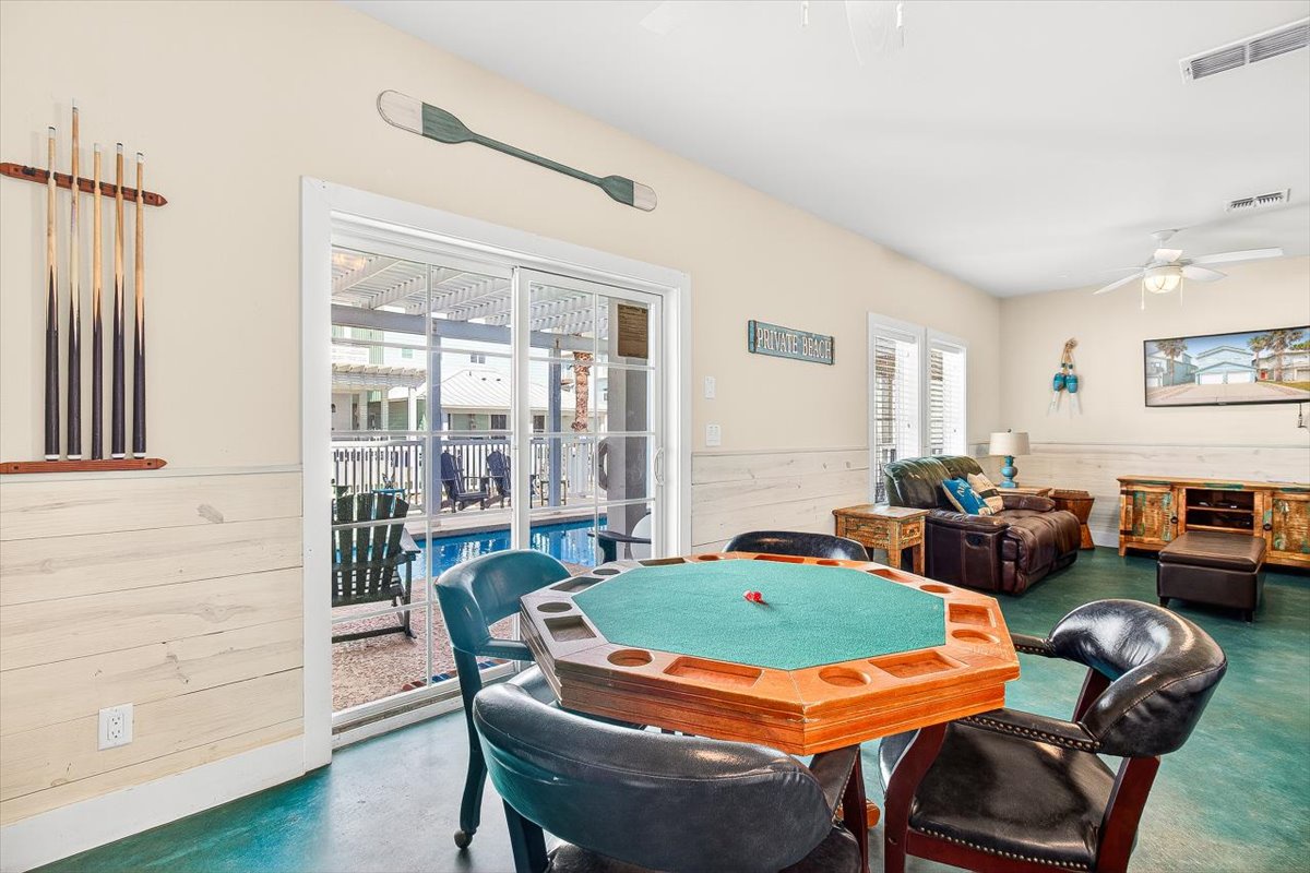 Your game room awaits with poker table, pool cues, and cozy seating overlooking the sparkling pool through glass doors.